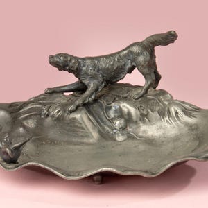 Op de afbeelding: Een asbak van zilverkleurig metaal met een gesculpteerd hondenbeeldje erop. De hond staat in een jachthouding, met zijn kop naar de zijkant gedraaid. De asbak heeft een gegolfde rand en een verhoogde basis.