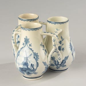 Op de afbeelding: Een set van drie crèmekleurige keramische kannen met blauwe handgeschilderde ontwerpen. Elke kan heeft een centraal paneel met een windmolen, omringd door bloemmotieven en een geschulpte blauwe rand langs de bovenkant en de voet.