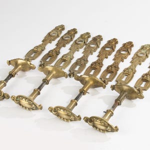 Puede incluir: Un conjunto de siete manijas de puerta antiguas de latón. Cada manija presenta un diseño ornamentado con un ojo de cerradura, un vástago decorativo y una base detallada. Las manijas están dispuestas en diagonal sobre un fondo blanco.