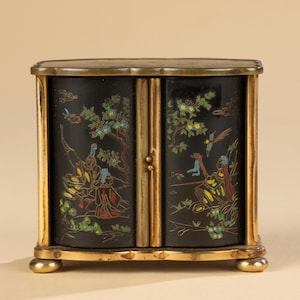 May include: A small, antique cabinet with a black front featuring a colorful Asian-inspired scene. The cabinet has gold-colored trim and small, rounded feet. The doors depict figures, trees, and birds.