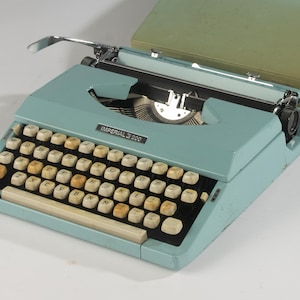 Puede incluir: Una máquina de escribir portátil Imperial 200 de color azul claro con teclado QWERTY y teclas de color blanco roto. La máquina incluye una palanca de retorno de carro de metal, un mecanismo de cinta negra y una tapa de estuche rígido verde oliva.