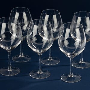 Può includere: Sei calici da vino trasparenti con steli lunghi e coppe arrotondate sono disposti su una superficie blu scuro. I calici sono vuoti e riflettono la luce, creando riflessi e ombre. I calici da vino sono eleganti.
