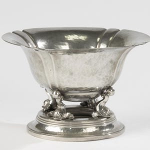 Op de afbeelding: Een zilverkleurige metalen kom met een gehamerde textuur. De kom heeft een geschulpte rand en wordt ondersteund door drie dolfijnvormige voetjes op een cirkelvormige basis. De kom is waarschijnlijk voor decoratieve of serveerdoeleinden.