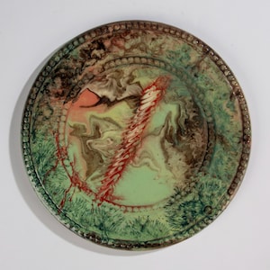 May include: A decorative ceramic plate with a unique design. The plate features a green and brown marbled pattern with a central red and white design. The rim has a beaded border and green accents, creating a textured appearance.