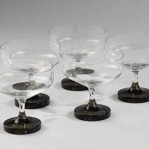 May include: Set of five clear glass champagne coupes with black bases. The coupes are arranged in a semi-circle on a white surface.
