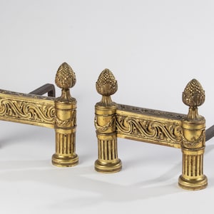 May include: A pair of gold-toned metal fireplace andirons featuring fluted column bases, ornate scrollwork detailing on the horizontal crossbars, and decorative finials shaped like pinecones at the top of each vertical post.
