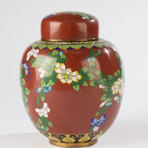May include: A red decorative jar with a lid, adorned with a floral pattern in white, yellow, and blue. The jar has a gold base and a detailed design on the body and lid. The overall design is ornate and detailed.