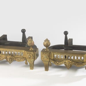 Pode incluir: Um par de morrões de lareira a condizer com frentes de metal dourado decoradas com motivos de cabeça de leão, festões e remates em forma de chama. Cada peça tem uma base retangular com quatro pernas e suportes de ferro preto.