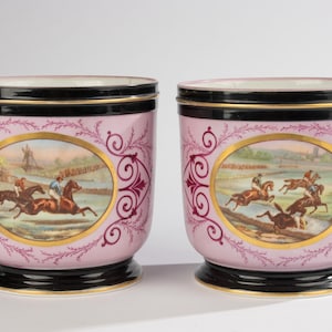 Puede incluir: Un par de maceteros cilíndricos de porcelana con cuerpo rosa claro y borde y base negros. Cada macetero muestra un medallón ovalado central con una escena pintada a mano de caballos y jinetes, enmarcada por adornos dorados y rojos oscuros.