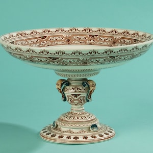 Può includere: Una ciotola in ceramica bianca con piedistallo e design floreale marrone e bianco. La ciotola ha una base larga e poco profonda e uno stelo alto e sottile con due maniglie decorative.