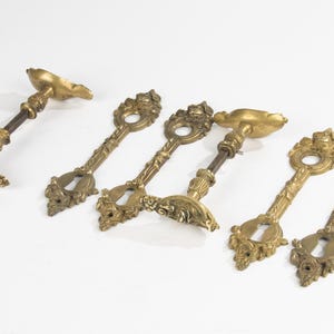 May include: A collection of antique brass door handles and keyhole covers. The handles have an ornate design with a gold finish. The keyhole covers are key-shaped, also with an ornate design. The items are arranged on a white surface.