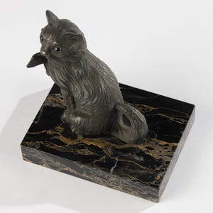 May include: A dark grey cat figurine with blue eyes sits on a black marble base with gold veining. The cat is sculpted with detailed fur and is posed in a seated position, looking to the side.