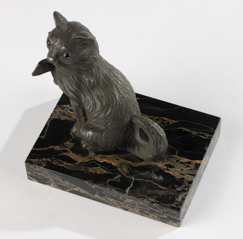 May include: A dark grey cat figurine with blue eyes sits on a black marble base with gold veining. The cat is sculpted with detailed fur and is posed in a seated position, looking to the side.