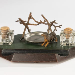 Puede incluir: Un tintero de escritorio con base hexagonal en verde oscuro y marrón, dos tinteros cuadrados de cristal transparente con tapones de corcho, una bandeja circular plateada y una figura de perro de metal color bronce junto a una estructura de ramas.