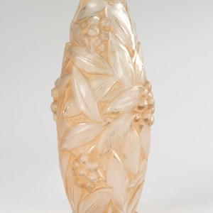 May include: A tall, peach-colored glass vase with a floral and leaf design. The vase has a wide base and a narrow neck, with the floral pattern covering the body. The design includes clusters of berries and long, slender leaves.