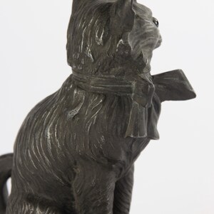 May include: A dark grey cat ornament, seated with a bow tie. The cat's fur is textured, and the figurine has a detailed, antique appearance. The cat is facing right, with its head turned slightly.
