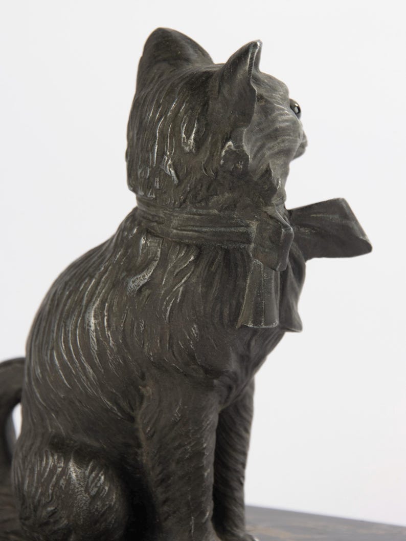May include: A dark grey cat ornament, seated with a bow tie. The cat's fur is textured, and the figurine has a detailed, antique appearance. The cat is facing right, with its head turned slightly.