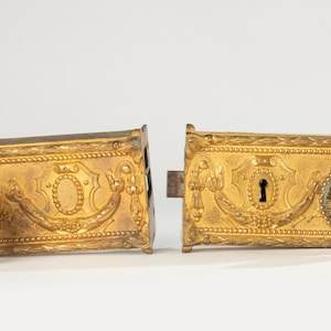 Puede incluir: Juego de cerradura de puerta dorada ornamentada. El juego se divide en dos piezas rectangulares, cada una con patrones de relieve detallados, incluyendo diseños florales y de volutas. Una pieza tiene un ojo de cerradura y un elemento circular decorativo.