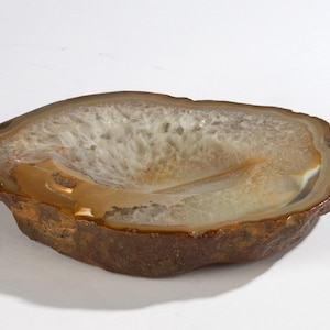 Op de afbeelding: Een natuurlijke agaatkom met een gepolijst interieur. De kom heeft een mix van bruine, witte en bruine kleuren, met een ruwe, getextureerde buitenkant. De agaat is in een afgeronde vorm gesneden, geschikt voor decoratief gebruik.