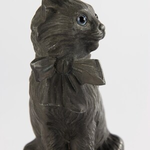 May include: A dark grey cat ornament with blue eyes and a bow tie. The cat is depicted in a seated position, with its head turned to the side. The ornament has a detailed texture, giving it a realistic appearance.