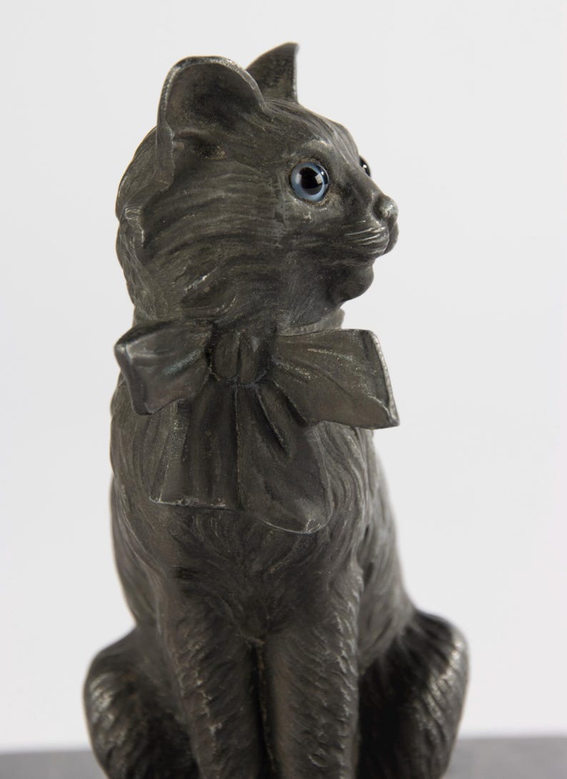 May include: A dark grey cat ornament with blue eyes and a bow tie. The cat is depicted in a seated position, with its head turned to the side. The ornament has a detailed texture, giving it a realistic appearance.