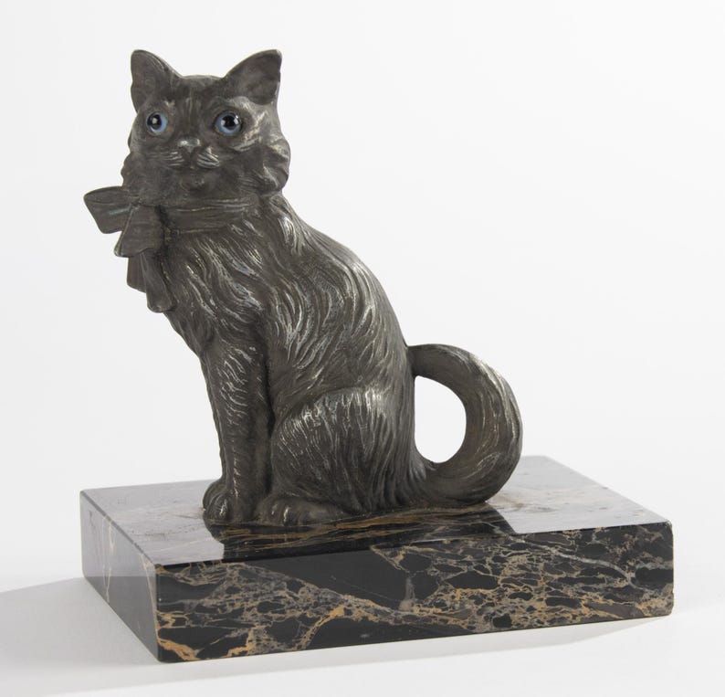 May include: A dark grey cat ornament with blue eyes and a bow, sitting on a black and gold marble base. The cat has detailed fur and a curled tail. This decorative sculpture is a unique home decor item.