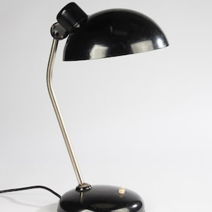 May include: A black desk lamp featuring a dome-shaped shade, a curved silver-toned metal neck, and a round black base. The lamp includes a small white push-button switch located on the top of the base and a black power cord extending from the rear.