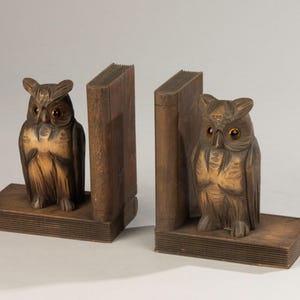以下が含まれることがあります： 木製のフクロウのブックエンドのペア。各ブックエンドは、本の山のようなベースに止まっている彫刻されたフクロウの姿を特徴としています。フクロウは詳細な羽と琥珀色の目をしています。ブックエンドは温かみのある茶色です。