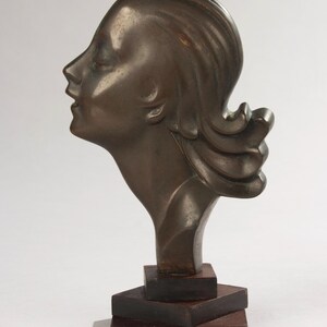 Könnte beinhalten: Bronzeskulptur eines Frauenprofils mit geschlossenen Augen und fließendem Haar. Die Skulptur ist auf einem dunklen Holzsockel montiert. Das Kunstwerk hat eine klassische, elegante Ästhetik.