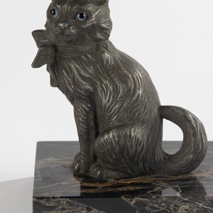 May include: A dark grey cat ornament with blue eyes and a bow tie, sitting on a black and gold marble base. The cat has detailed fur and a curled tail, creating a decorative sculpture.