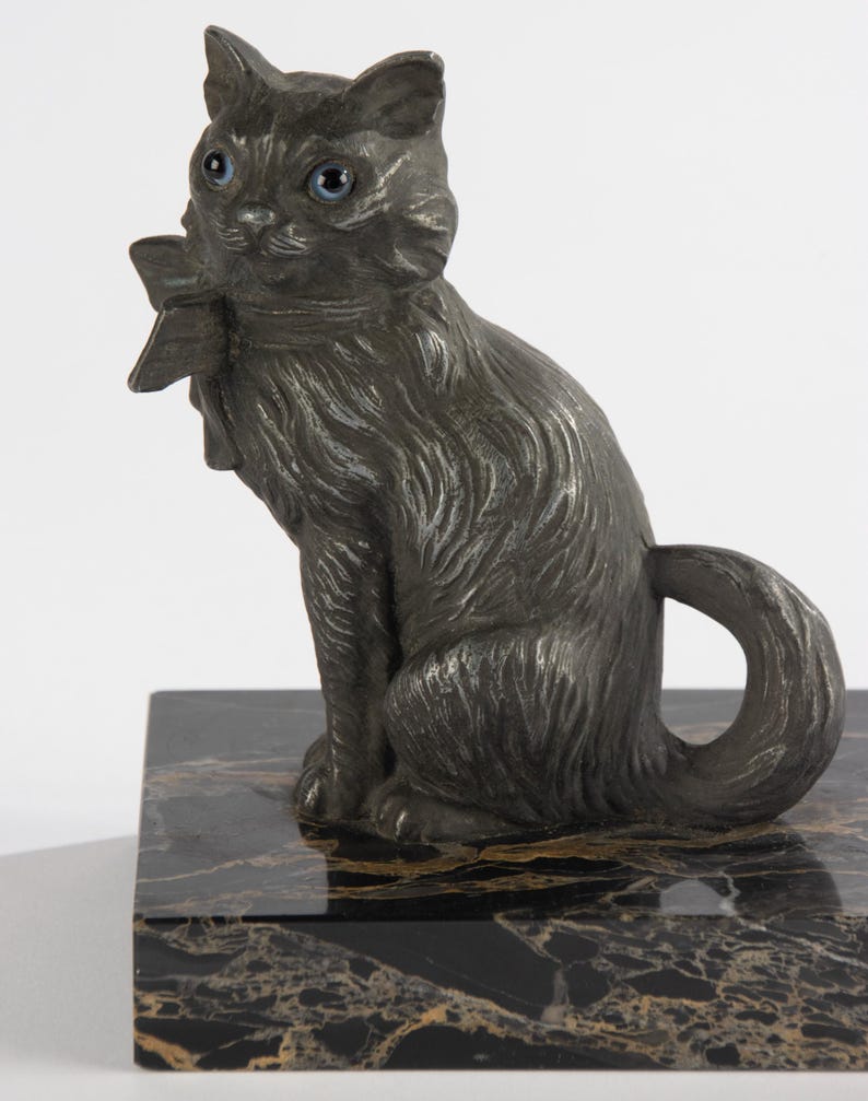 May include: A dark grey cat ornament with blue eyes and a bow tie, sitting on a black and gold marble base. The cat has detailed fur and a curled tail, creating a decorative sculpture.