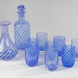 May include: A glass drinkware set consisting of one decanter with a spherical stopper, one wide-base carafe, and six matching tumblers. Each piece is made of clear glass featuring a distinct blue and white diagonal swirl pattern throughout.