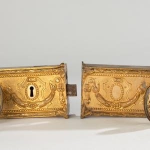 Peut inclure: Deux serrures de porte dorées ornées avec des détails complexes. Chaque serrure présente une poignée ovale et un trou de serrure. Les serrures sont probablement antiques ou vintage, avec un design décoratif.