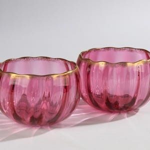 Op de afbeelding: Twee roze glazen kommen met een geschulpte rand en gouden rand. De kommen hebben verticale ribbels en staan tegen een witte achtergrond. De kommen zijn waarschijnlijk decoratief of om te serveren.