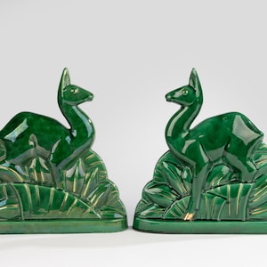 Może przedstawiać: Para szmaragdowozielonych ceramicznych podpórek do książek przedstawiających stylizowaną figurę jelenia spoczywającą na wachlarzowatej podstawie z liści. Błyszczące wykończenie podkreśla gładkie kontury zwierzęcia oraz promieniste wzory liści.