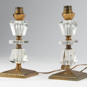 Op de afbeelding: Twee heldere kristallen tafellampen met messing voetstukken en accenten. De lampen hebben een geometrisch ontwerp met facetgeslepen kristallen kappen en vierkante voetstukken. De lampen zijn verbonden door een bruin elektrisch snoer.