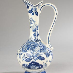 Peut inclure: Une cruche en céramique blanche ornée d'un motif floral bleu. La cruche a un col fin, une anse incurvée et un bec évasé. Le motif floral bleu comprend des fleurs et des feuilles. La base est décorée de rayures bleues. Hauteur d'environ 30 cm.