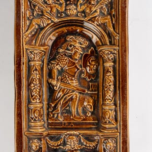 Op de afbeelding: Een rechthoekige, bruine keramische plaat met een reliëfsculptuur die een krijgersfiguur afbeeldt die een zwaard en een afgehakt hoofd vasthoudt. De plaat heeft sierlijke architectonische details en cherubijnse figuren.