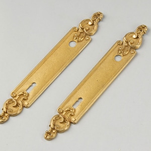 Może przedstawiać: Dwie złote płytki drzwiowe z ozdobnymi, zwiniętymi wzorami u góry i na dole. Każda płytka ma dziurkę od klucza i okrągły otwór.