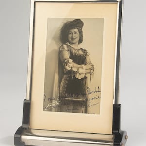 May include: A rectangular silver-toned metal picture frame with black accents, holding a sepia-toned photograph of a person wearing a feathered hat and a detailed costume. The photograph features handwritten cursive text in black ink across the bottom.