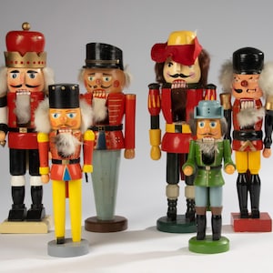 May include: A collection of six wooden nutcrackers in various sizes and designs. The nutcrackers feature vibrant colors, including red, black, yellow, and green, with detailed facial features and traditional military-style uniforms. They stand on individual bases.