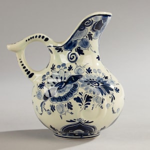 Może przedstawiać: Ceramiczny dzbanek w kremowym kolorze z misternymi niebieskimi wzorami kwiatowymi. Dzbanek posiada zakrzywiony uchwyt z kropkowanymi akcentami i dzióbek. Projekt nawiązuje do tradycyjnej ceramiki z Delft. Wysokość ok. 20 cm.