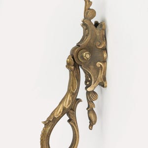 Puede incluir: Aldaba de puerta de latón ornamentada con un diseño floral detallado. La aldaba presenta un asa curvada y una placa trasera decorativa, montada sobre una superficie blanca. El latón tiene una pátina cálida y envejecida.