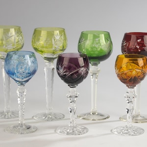 May include: A collection of seven multi-colored crystal wine glasses featuring intricate cut-glass patterns on the bowls and clear, faceted stems. The set includes glasses in shades of light green, blue, dark green, purple, amber, and deep red.
