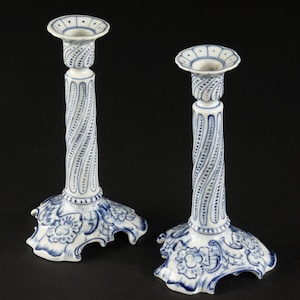 May include: A pair of white ceramic candlesticks featuring intricate blue hand-painted floral patterns on the wide, scalloped bases and spiralling vertical columns with dotted detailing leading up to the circular candle holders at the top.