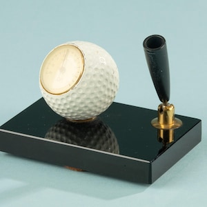 Op de afbeelding: Een golf-thema bureauset met een witte golfbal klok met een goudkleurige rand en een zwarte pen. De klok en penhouder zijn gemonteerd op een zwarte rechthoekige basis. De pen is zwart met een goudkleurige accent.