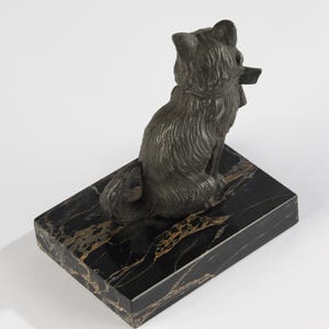 May include: A bronze-coloured cat figurine seated on a rectangular black marble base with gold veining. The cat is facing away, holding a small object in its mouth. The sculpture has detailed fur and a collar.