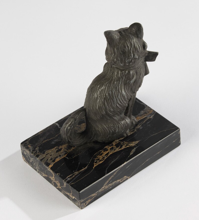 May include: A bronze-coloured cat figurine seated on a rectangular black marble base with gold veining. The cat is facing away, holding a small object in its mouth. The sculpture has detailed fur and a collar.