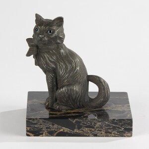 May include: A dark grey cat ornament with blue eyes and a bow, perched on a black and gold marble base. The cat features detailed fur and a curled tail, a decorative sculpture.