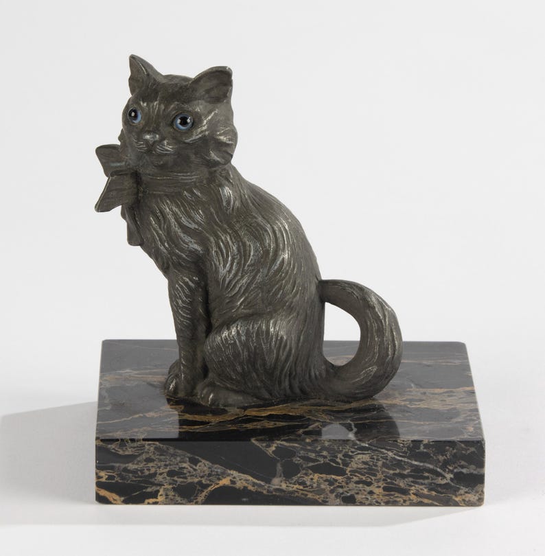 May include: A dark grey cat ornament with blue eyes and a bow, perched on a black and gold marble base. The cat features detailed fur and a curled tail, a decorative sculpture.